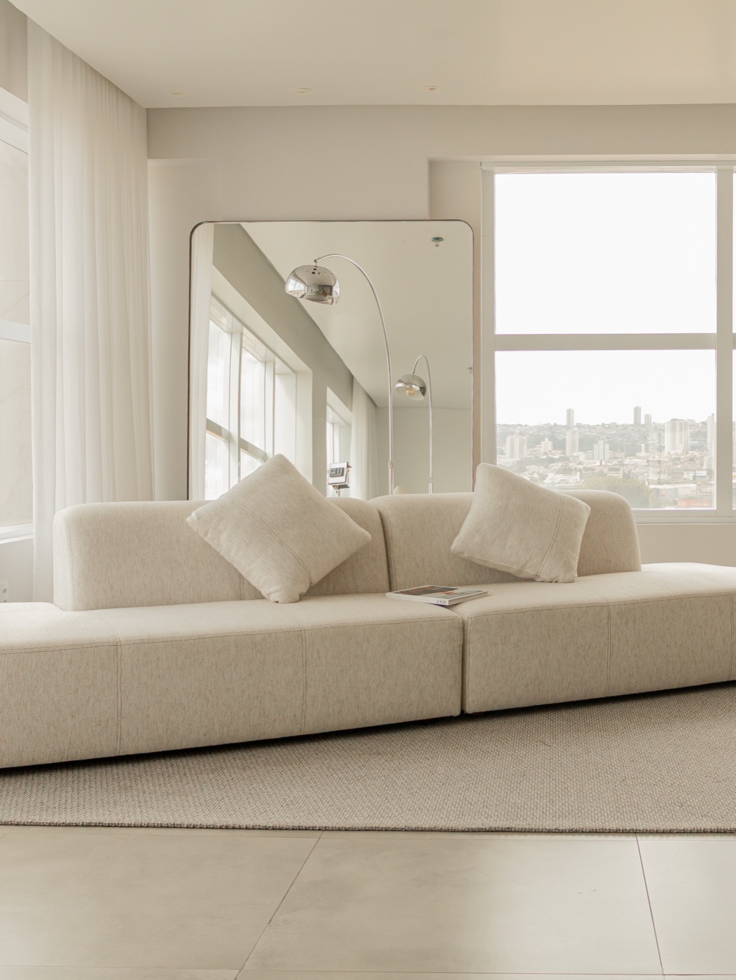 Sofá modular creme diante do espelho de corpo inteiro e das cortinas difusoras
